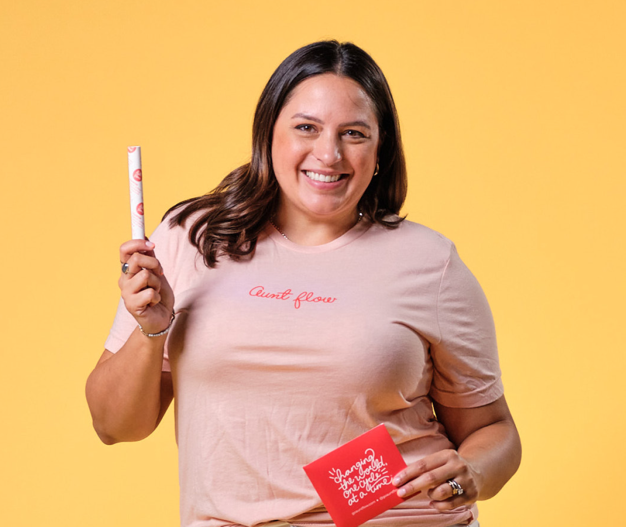 Model wearing Aunt Flow "Changing the World, One Cycle at a Time" peach colored t-shirt. Posing with Aunt Flow Cartridge Tampon and Pad